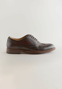 Chaussure Oxford en cuir brun foncé à bout golf avec détails brogue, laçage fermé et talon empilé sur fond clair.
