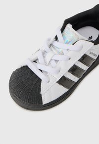 Baskets noir et blanc avec un embout texturé, des lacets blancs et trois bandes noires sur le côté. Comprend une étiquette de marque irisée.