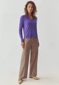 Maglione lavorato a maglia viola con scollo a V, abbinato a pantaloni beige a gamba larga e scarpe basse di colore chiaro. Texture liscia, design casual.