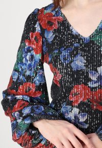 Blouse noire à fleurs avec des fleurs rouges et bleues, texture pailletée, manches bouffantes et décolleté en V.