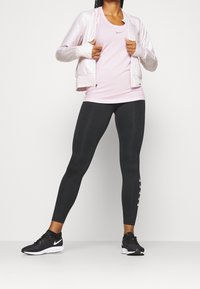 Ljusrosa zip-up jacka över en matchande tanktop, svarta leggings med vit logodetalj och svarta träningsskor.