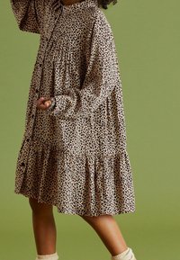 Leopardikuvioinen mekko, joka on valmistettu rennosta kankaasta. Mekossa on nappisuljenta, pitkät hihat ja kerroksellinen helma. Väri: beige tummilla laikkuilla.