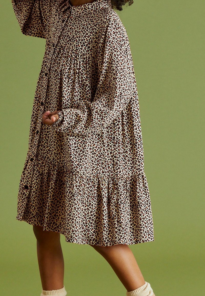 Leopardikuvioinen mekko, joka on valmistettu rennosta kankaasta. Mekossa on nappisuljenta, pitkät hihat ja kerroksellinen helma. Väri: beige tummilla laikkuilla.