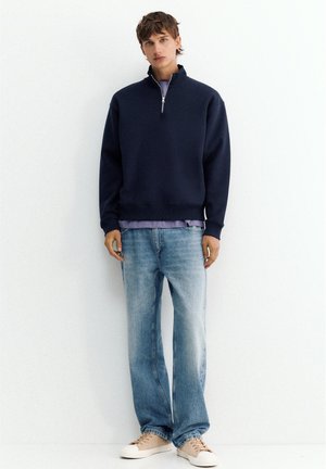 Jeune homme debout portant un pull bleu marine à fermeture éclair quart, chemise violet clair en dessous, jean bleu ample et baskets beiges contre un mur blanc.
