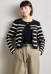 Cardigan a righe bianche e nere con scollatura rotonda, chiusura frontale con bottoni e texture a coste, abbinato a pantaloni khaki a vita alta.