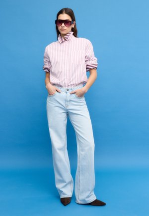 Kobieta w oversize'owych jasnoniebieskich dżinsach, pasiastym różowym swetrze z podwiniętymi rękawami, ciemnych, szpiczastych butach i dużych fioletowych okularach przeciwsłonecznych stoi na tle niebieskiego tła.