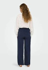 Blouse en dentelle blanche avec un motif festonné, associée à un pantalon large bleu marine avec une taille élastique. Des sandales marron clair sont visibles.