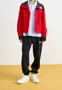 Rote wasserdichte Jacke mit schwarzen Akzenten, darüber ein hellblaues Hemd. Schwarze Jogginghose und weiße Sneakers vervollständigen das Outfit.