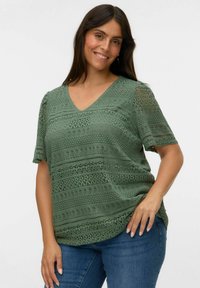 Femme souriante avec de longs cheveux noirs, portant un haut vert en dentelle à manches courtes et en V, ainsi que des jeans bleus, posant avec une main sur la hanche.