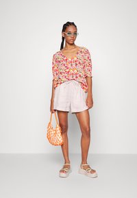 Blusa com estampado floral e mangas curtas, calções de cordão em bege claro, sandálias verdes e uma bolsa de rede laranja. Modelo em pé sobre um fundo simples.