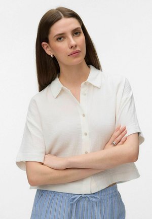 Femme aux longs cheveux bruns, portant une chemise blanche à manches courtes boutonnée et un pantalon rayé bleu, debout les bras croisés.