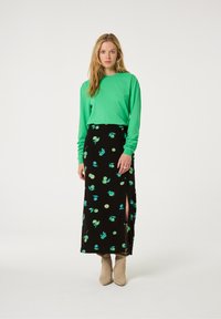 Groene blouse met lange mouwen, gecombineerd met een zwarte bloemenrok, voorzien van groene en blauwe patronen, zijsplitsingen en beige enkellaarsjes.