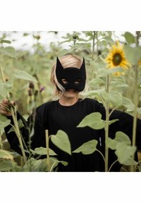 Svart filtmask med spetsiga öron, bärs av ett barn i en lös svart dräkt, omgiven av gröna solrosor och höga blad.