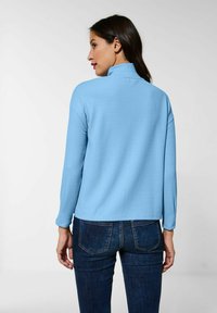 Pullover azzurro chiaro, strutturato, con colletto alto e maniche lunghe, abbinato a jeans scuri. Presenta una vestibilità rilassata e un taglio di lunghezza media.