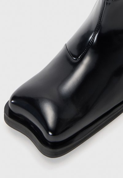 Black leather ankle boot with a glossy finish, rounded toe, and a low rubber sole. Seam detailing along the upper for structure.