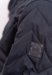 Quilted black puffer jacket detail featuring a textured square badge with circular design and contrasting stitching on smooth fabric.