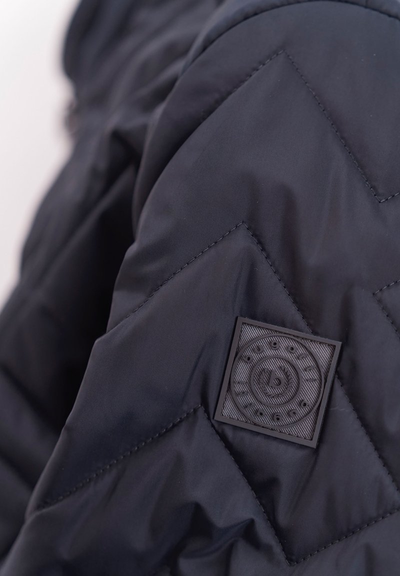 Gepolsterte schwarze Steppjacke mit Detail eines strukturierten quadratischen Abzeichens mit kreisförmigem Design und kontrastierenden Nähten auf glatter Oberfläche.