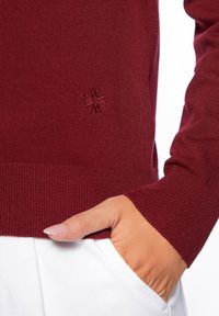 Maglione in maglia borgogna con polsini e orlo a coste. Presenta un logo ricamato discreto sul petto. Texture liscia, design aderente.
