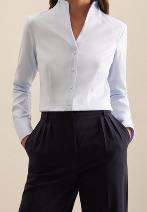 Vrouw met een lichtblauw overhemd met knopen, ingestopt in hoge, geplooide zwarte broek, met handen in de zakken.