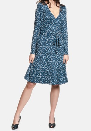 Kobieta ubrana w niebieską, kwiecistą sukienkę kopertową sięgającą do kolan, z długimi rękawami, dekoltem w kształcie litery V, koronkowym detalem na ramionach oraz czarnymi, otwartymi na palce szpilkami.