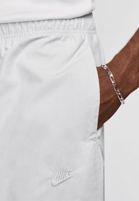 Os calções atléticos brancos apresentam uma cintura elástica, bolsos laterais e um logótipo da Nike bordado na perna esquerda. Uma pulseira de corrente prateada é visível.