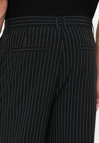 Pantalon noir à fines rayures avec passants de ceinture et deux poches passepoilées au dos, porté avec un haut noir texturé.