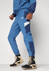 Blauwe sportbroek met contrasterende marineblauwe en witte panelen, elastische boorden en een logo op de bovenbeen, gecombineerd met zwart en tan hoge sneakers.