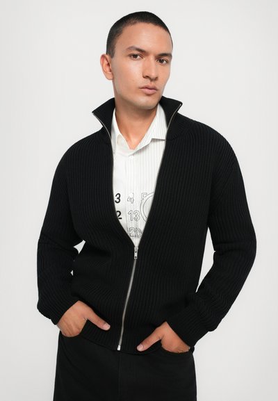 Black ribbed zip-up cardigan with high collar. Worn over a white striped shirt featuring a graphic print. Model poses with hands in pockets.