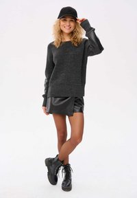 Schwarzer Strickpullover mit strukturierten Ärmeln, kombiniert mit einem schwarzen Leder-Minirock und schnürbaren Kampfstiefeln. Das Modell trägt eine schwarze Mütze.