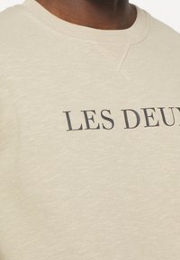 Sudadera beige de cuello redondo con el texto negro "LES DEUX" parcialmente visible en el pecho, llevada por una persona de piel oscura.