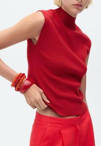 Haut rouge sans manches avec un col haut, en tissu lisse. Le modèle porte plusieurs bracelets en caoutchouc rouge vif, assortis à un pantalon rouge.