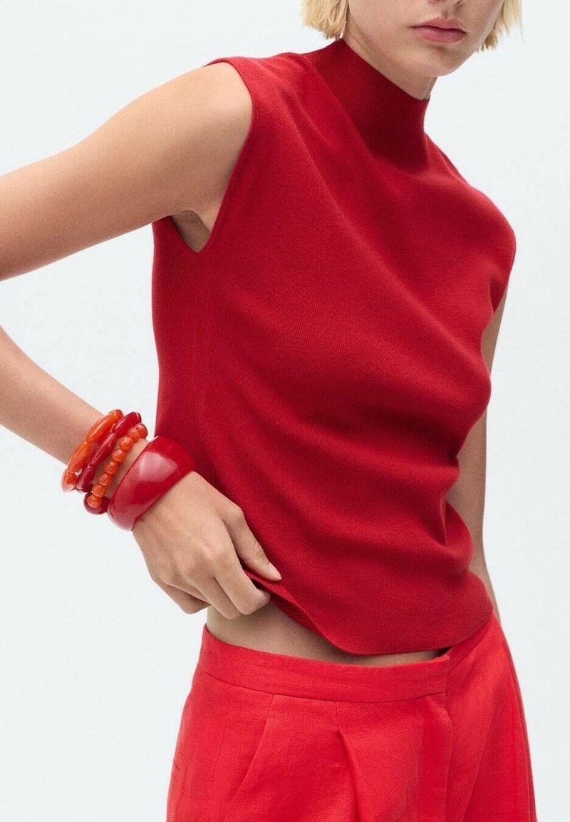 Haut rouge sans manches avec un col haut, en tissu lisse. Le modèle porte plusieurs bracelets en caoutchouc rouge vif, assortis à un pantalon rouge.