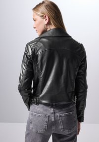 Giubbotto in pelle nera con spalle trapuntate, design cropped e dettagli in metallo argentato. Indossato su jeans denim grigi, evidenziando una silhouette elegante.