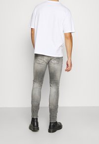 Muž v bledě šedých vybledlých skinny džínách, černých kožených botách a jednoduchém bílém tričku s krátkým rukávem, stojící čelem k odvrácené straně.