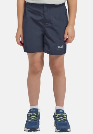 Kind draagt marineblauwe Jack Wolfskin short en blauwe wandelschoenen met witte sokken, staand tegen een effen achtergrond.