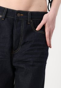 Temno modri jeans imajo prilagojeno zasnovo z rumenim šivanjem in zaponko. Žepi so vidni na sprednji strani.
