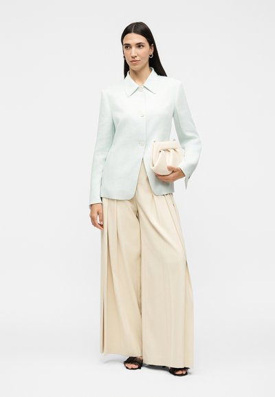 Γυναίκα φοράει γαλάζιο σακάκι με κουμπιά και φαρδιές μπεζ παντελόνες, κρατώντας μια κρεμ τσάντα φάκελο με μαύρα πέδιλα με ανοιχτά δάχτυλα σε απλό φόντο.