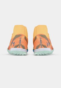 Pár oranžových a černých vysokých fotbalových bot s světle zelenými podrážkami, z pohledu zezadu na bílém pozadí.