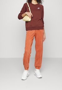 Kvinna iklädd en brun Nike-tröja, orange Nike-joggingbyxor, vita sneakers och håller en ljusgul axelväska framför en neutral bakgrund.