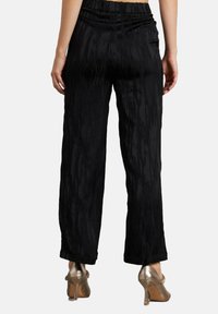 Vrouw in zwarte gestreepte wijde pijpen broek en metallic hoge hakken, van achteren getoond op een effen achtergrond.