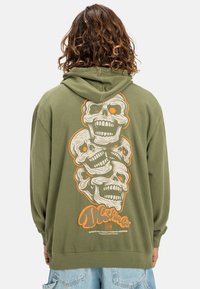 Felpa verde oliva con un grafiche di teschi impilati in bianco e arancione sul retro. Realizzata in tessuto morbido con polsini e orlo a costine.
