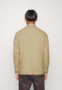 Camicia a maniche lunghe di colore marrone chiaro, realizzata in twill di cotone, dotata di colletto, polsini con bottoni e una vestibilità rilassata, mostrata da dietro.