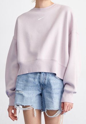 Personne portant un sweat-shirt à manches longues violet clair avec un petit logo blanc et un short en jean bleu clair usé.