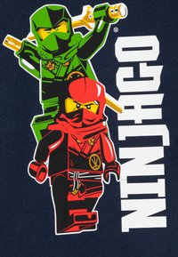 Lego Ninjago-grafikken har to karakterer i grønn og rød. Bakgrunnen er marineblå, med "NINJAGO" i fete hvite bokstaver.