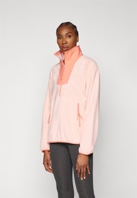Fleece pullover vaaleanpunaisena, jossa on korkea kaulus ja puolivetoketju. Siinä on oranssit yksityiskohdat ja vetoketjulliset taskut. Rakenteellinen, pehmeä kangas.