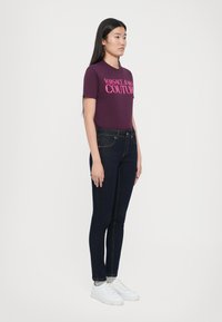 Lila rövidujjú póló, amelyen rózsaszín "Versace Jeans Couture" felirat található, sötétkék skinny farmernadrággal és fehér sportcipővel párosítva.