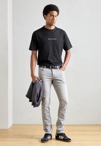Zwarte katoenen t-shirt met wit logo, lichtgrijze spijkerbroek, grijze sweatshirt over de arm, zwarte sneakers met witte strepen, casual fit.