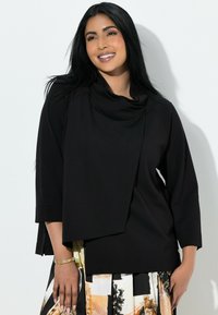 Czarny top z drapowanym kapturem i rękawami do połowy łokcia; posiada warstwowy fason o gładkiej fakturze. Akcenty stanowi złota bransoletka.