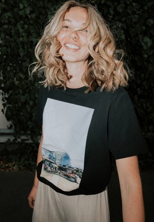 Zwarte T-shirt met een grote fotografische print van een modern gebouw en tuin. Model draagt lichte, vloeiende broeken; haar is golvend.