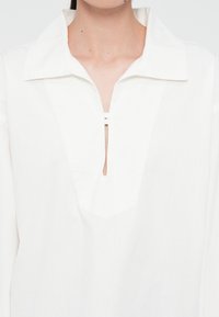 Biała koszula z kołnierzykiem, częściowym listwą na guziki i otworem w kształcie klucza na środku. Wykonana z lekkiego materiału o gładkiej teksturze.